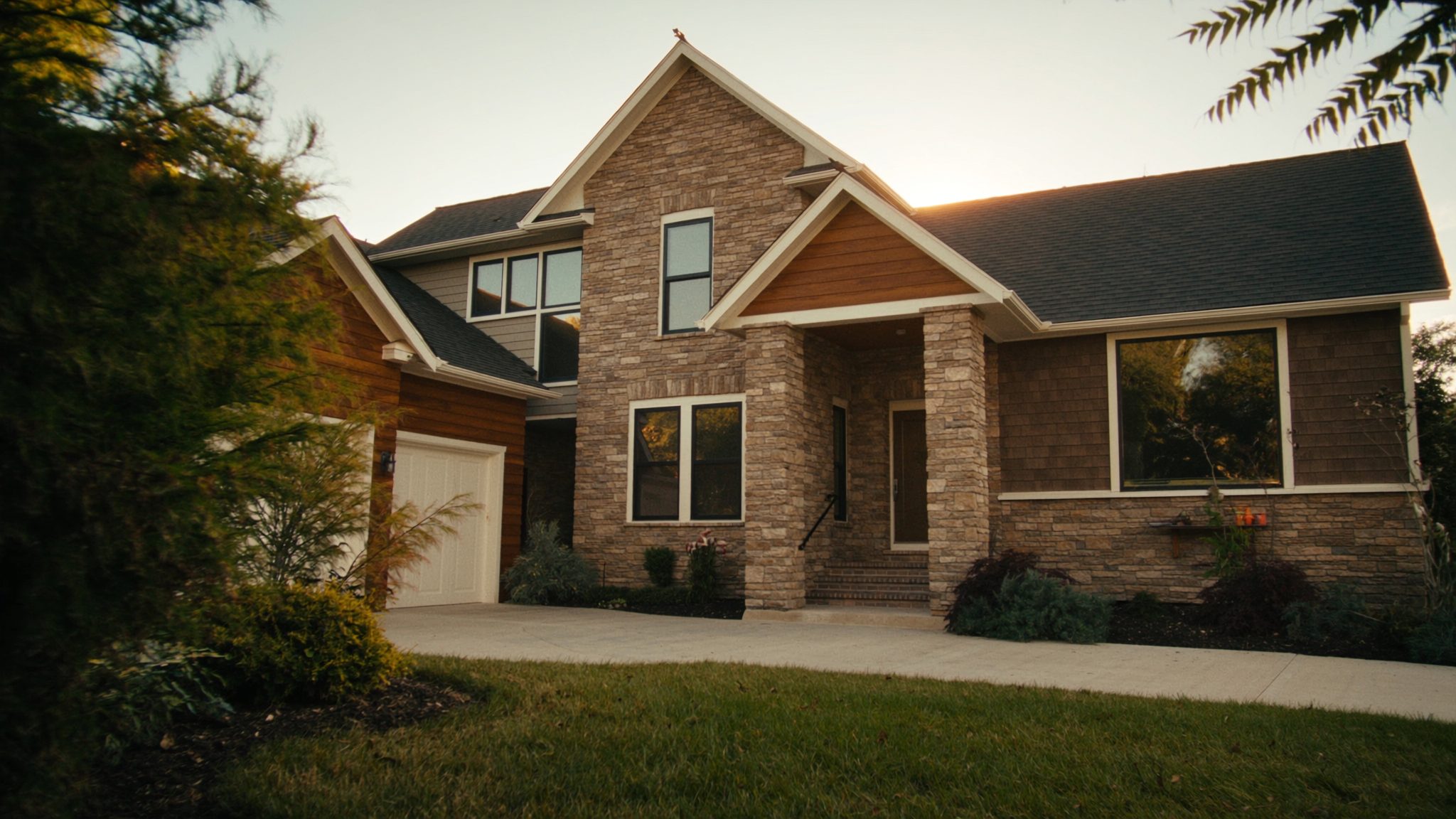 A modern two-storey home with stone and timber detailing at golden hour, framed by a leafy garden